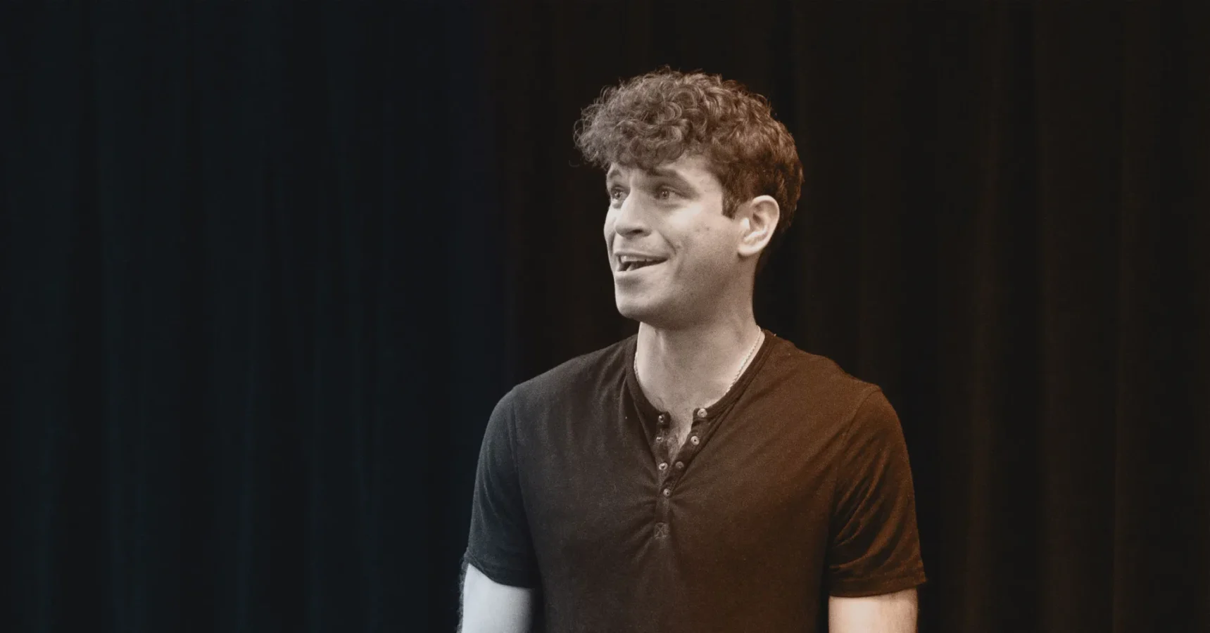 A young man with curly hair stands in front of a dark background, wearing a short-sleeve henley shirt and smiling slightly.