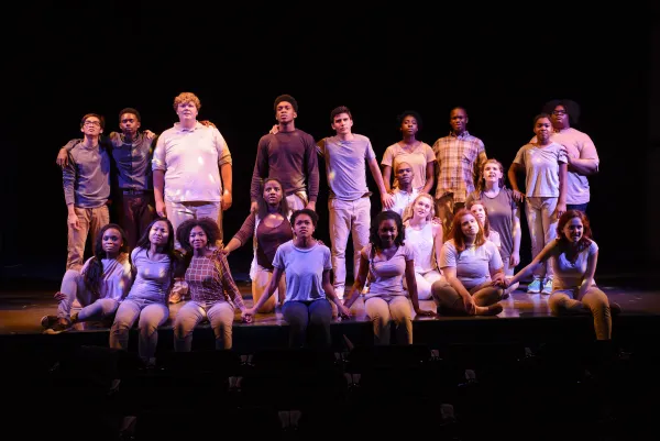 A group of young people stand and kneel on stage under dramatic lighting, facing forward, as if performing in a theater production.