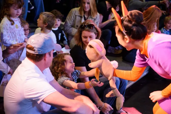 A performer in costume holds out a stuffed animal to a smiling child in an audience, with several adults and children seated around them.