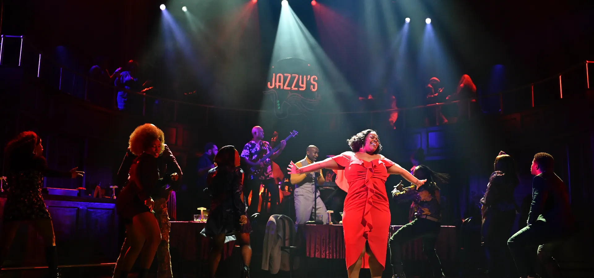 Performers sing and dance energetically on a stage set resembling a jazz club called "Jazzy's," with vibrant lighting and a live band in the background.