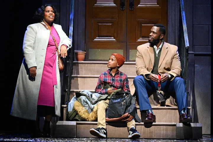 Three people sit on brownstone steps, with one woman standing and talking. The man and a child with a backpack sit, all dressed in winter clothes, in front of a wooden door.