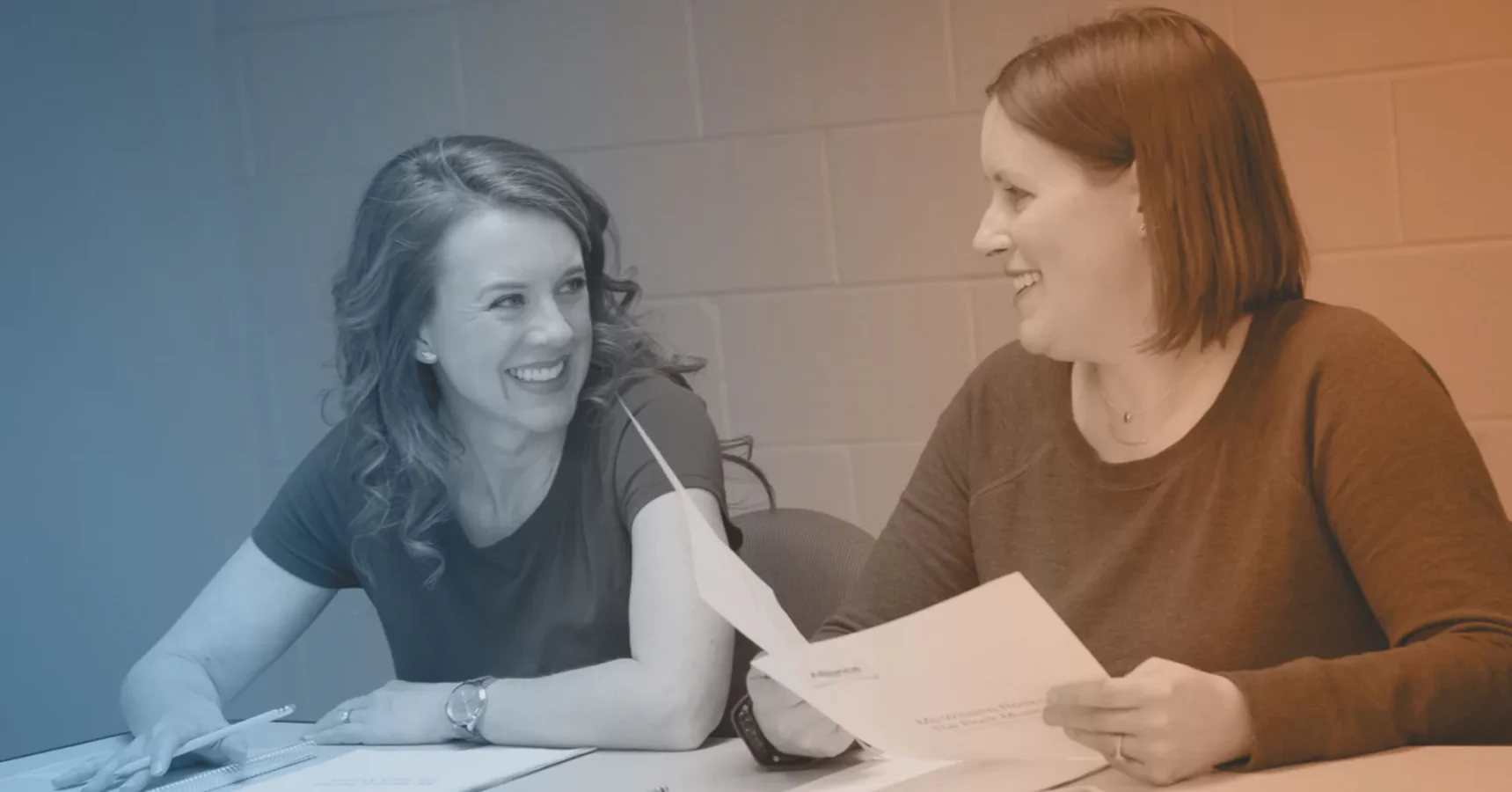 Two women sit at a table, holding papers and smiling at each other in a discussion.