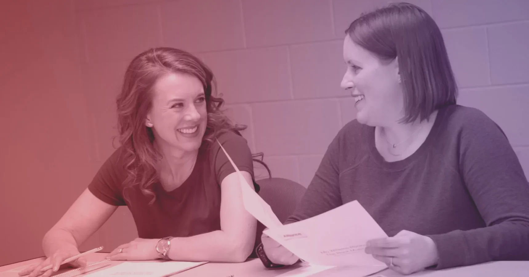 Two women sitting at a table, smiling and talking while looking at papers in their hands.