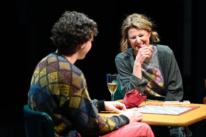 Two people sit at a table playing Scrabble, with a glass of white wine and a red bag on the table. One person is smiling and the other is facing away from the camera.