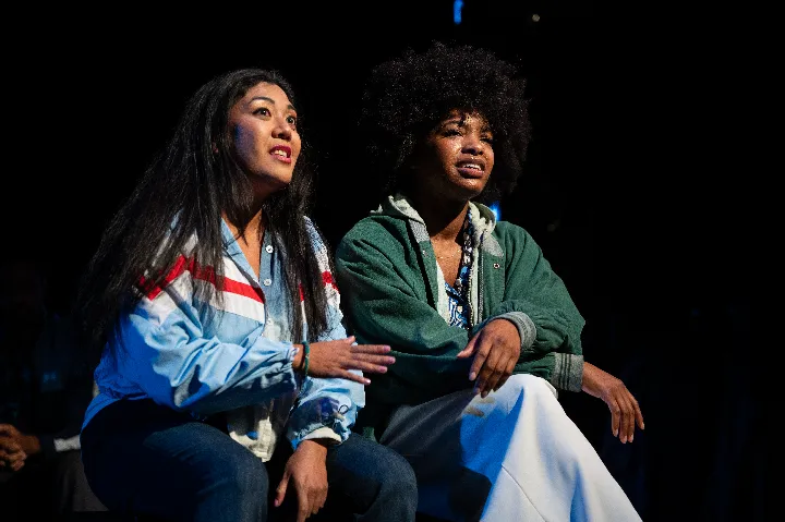 Two people sit close together on stage, illuminated by a spotlight, wearing casual clothing and looking slightly upward with engaged expressions.