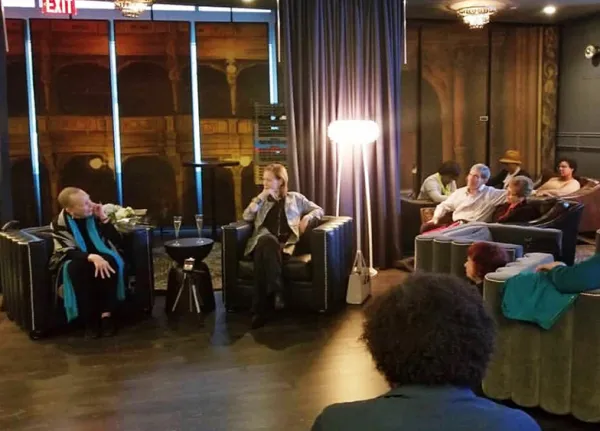 Several people are seated in a dimly lit lounge, listening to a speaker with a microphone during what appears to be a small, informal event.