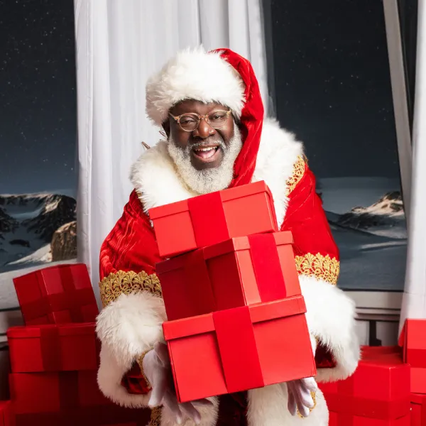 A man dressed as Santa Claus holds three stacked red gift boxes, with more red boxes in the background and a snowy landscape visible through the window.
