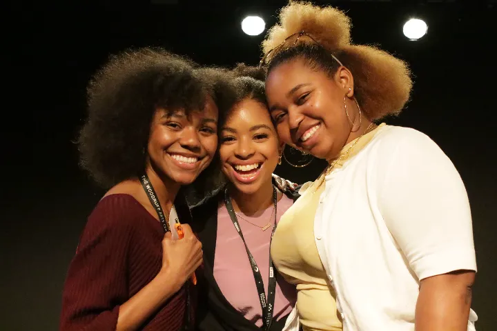 Three young women stand close together, smiling at the camera, wearing event lanyards with a dark background behind them.