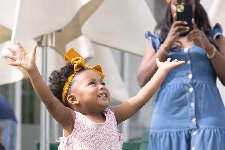 A young girl with a yellow headband smiles and raises her arms while a woman in a blue dress takes a photo with her phone in the background.