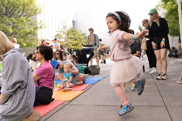 A young girl in a tutu jumps in the air while people, including children and adults, sit and watch on colorful mats at an outdoor event.