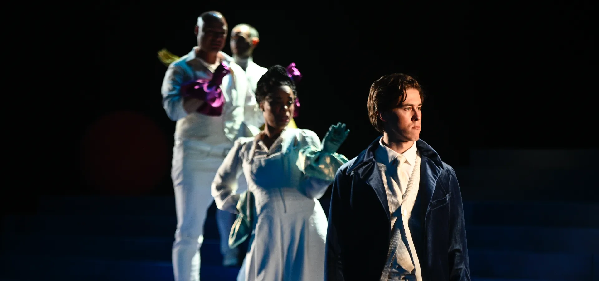 Four people in period costumes stand on stage under dramatic lighting; the person in front wears a dark coat and looks away while the others stand behind in white attire with colorful gloves.