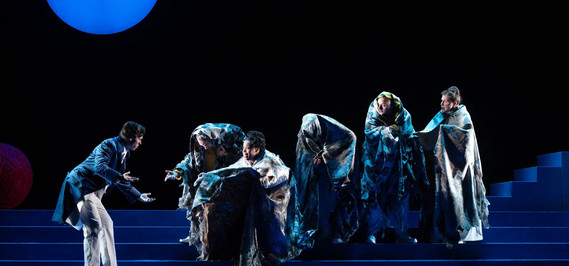 A person in a suit gestures toward five people draped in patterned cloaks on blue-lit steps, performing on a dark stage under a large blue circle.