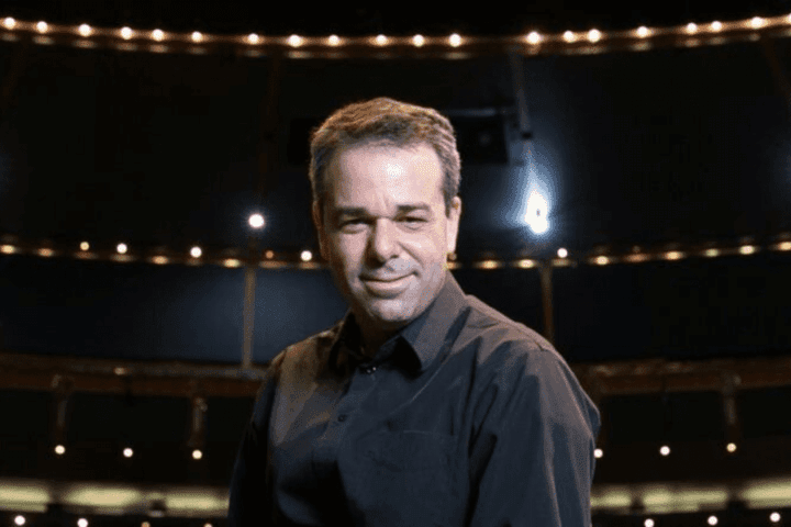 A man in a dark shirt stands under stage lights with an auditorium in the background, looking toward the camera.