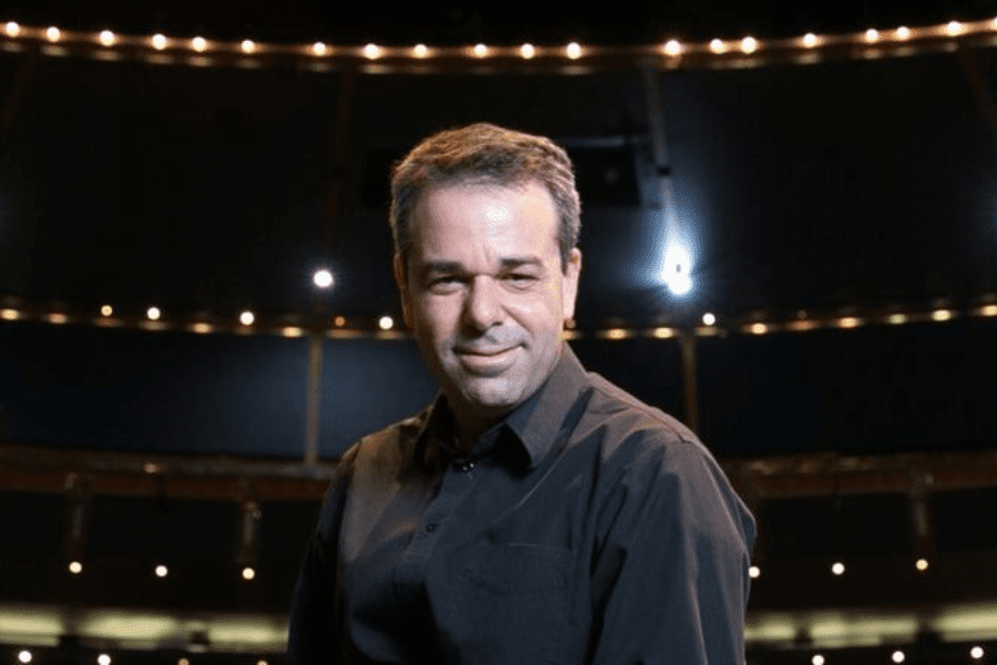 A man in a dark shirt stands under stage lights with an auditorium in the background, looking toward the camera.