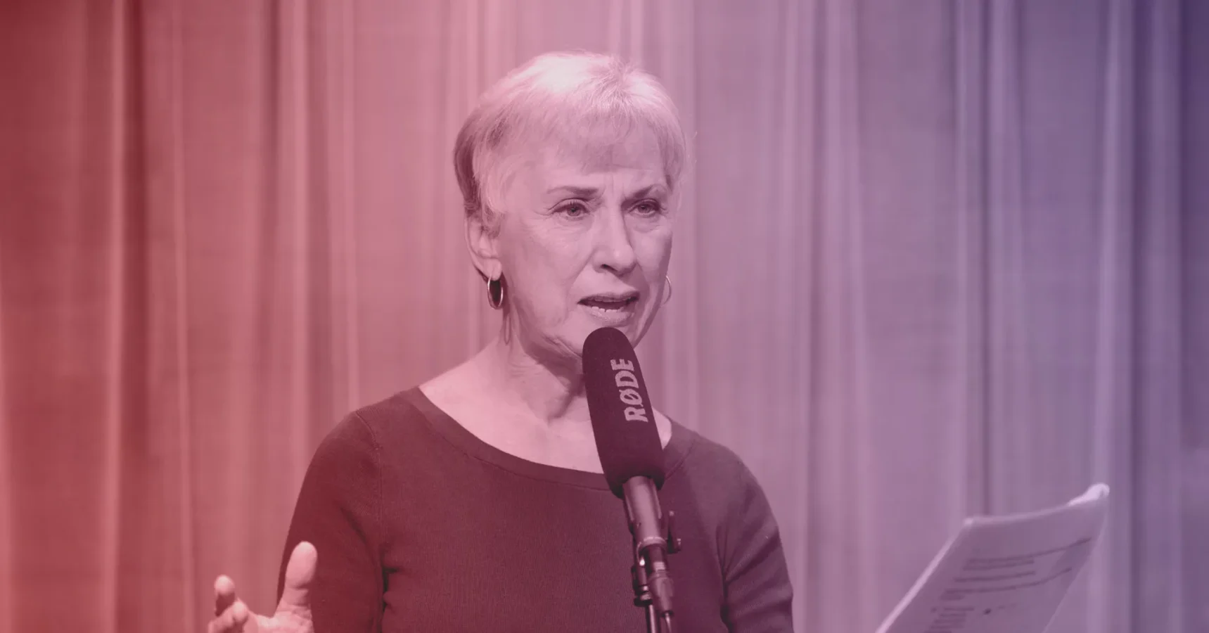 An older woman with short gray hair speaks into a RODE microphone while holding papers, standing in front of a curtain backdrop.