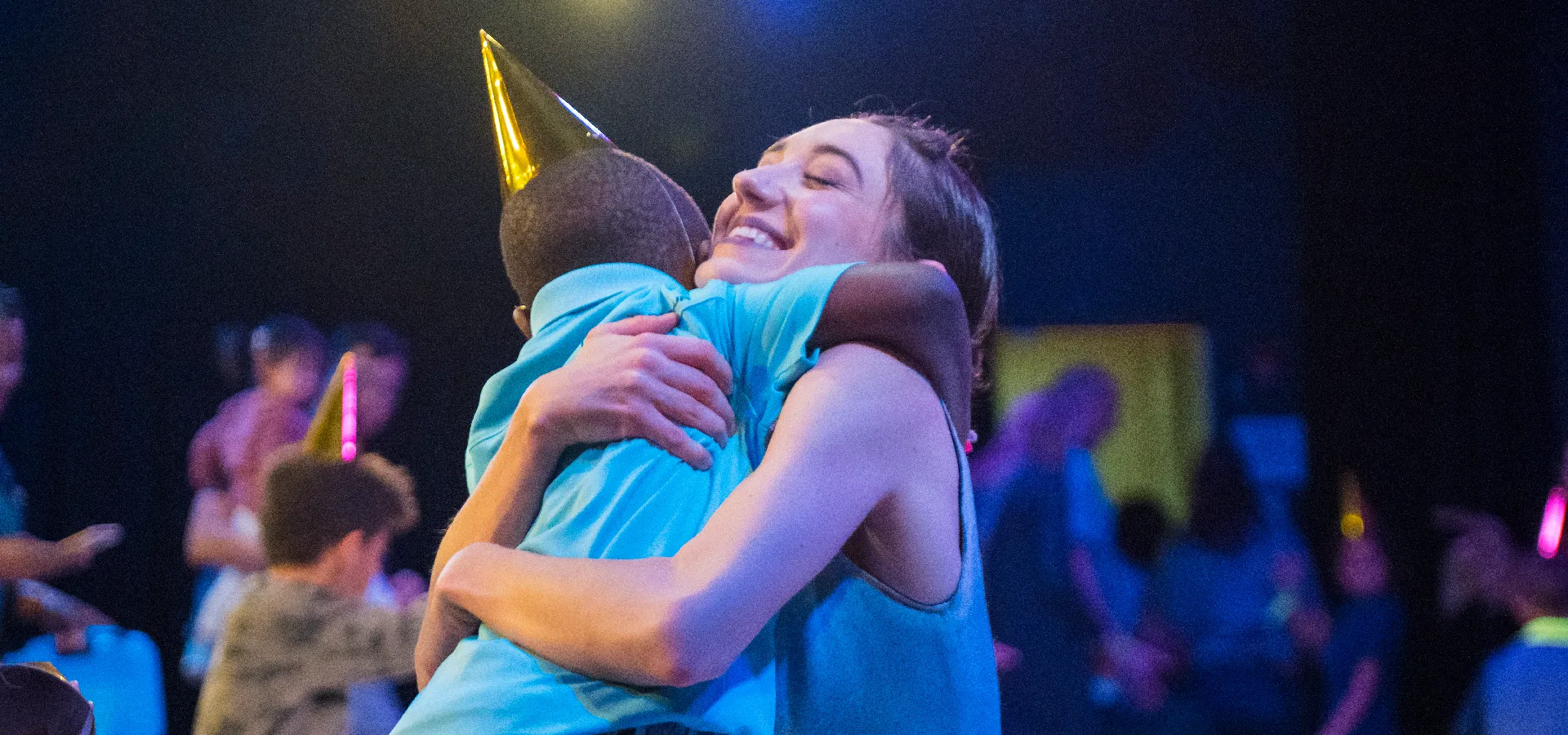 A woman smiling and hugging a child wearing a party hat at a celebration, with other people and party hats visible in the background.