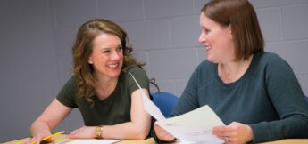 Two women sit at a table in a meeting room, holding papers and smiling at each other during a discussion.
