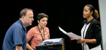 Three people are gathered around a piano, with one woman holding sheet music and singing while the other two listen attentively.
