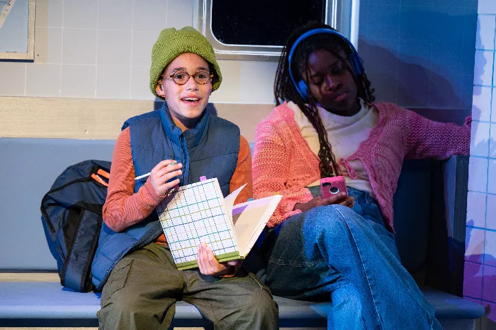 Two people sit on a bench; one is smiling and holding an open book with a grid while the other, wearing headphones, looks at a phone.