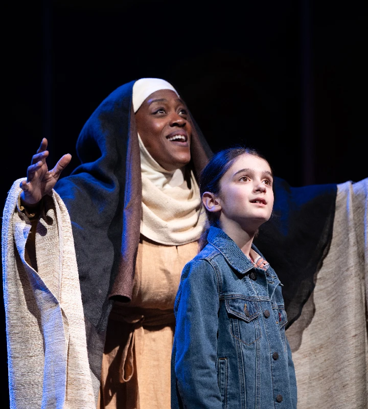 An adult in biblical costume stands with arms raised behind a young girl in a denim jacket, both looking upward under stage lighting.