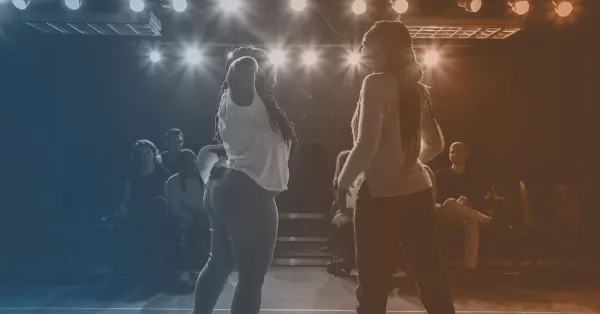 Two people dance on a stage under bright lights, with an audience seated in the background.