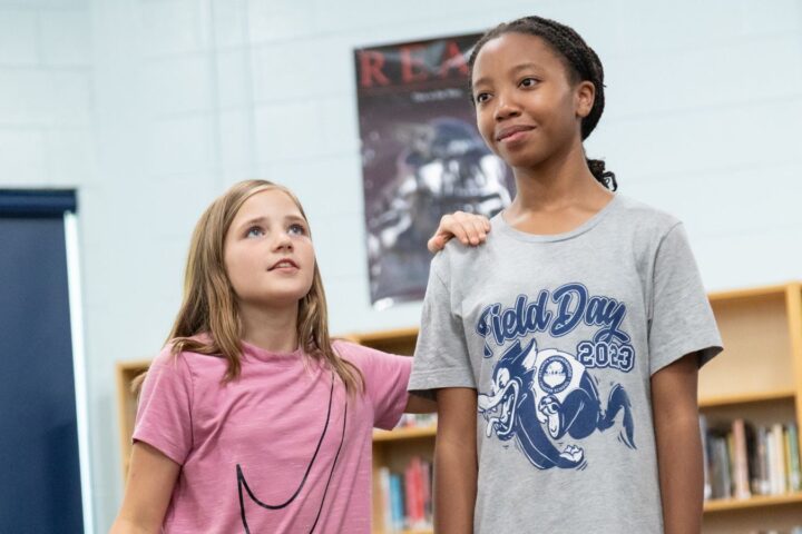 Two girls stand in a library; one with her hand on the other's shoulder. Bookshelves and posters are visible in the background.