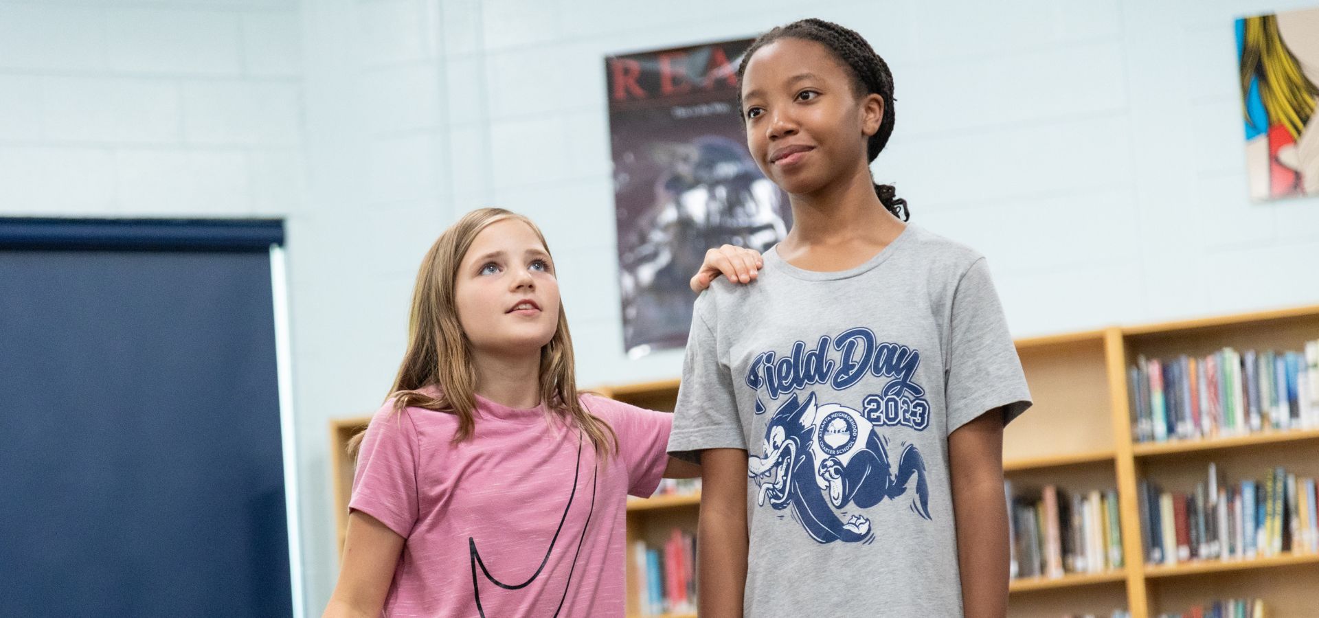 Two girls stand in a library; one with her hand on the other's shoulder. Bookshelves and posters are visible in the background.