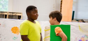 Two boys stand facing each other indoors in acting camp; one wears a plain yellow shirt, the other wears a green costume. Drawings are displayed on the wall behind them.