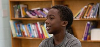 A boy in a gray shirt sits and smiles in a room with bookshelves filled with books in the background at acting camp.