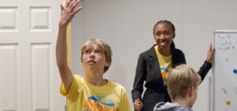 A boy raises his hand while two other kids, one standing by a whiteboard in acting camp. All wear yellow t-shirts with a sun graphic.