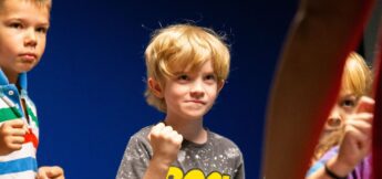 Three young children stand indoors, two with fists raised as if practicing a martial arts or boxing stance, against a solid blue background.