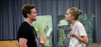 Two people stand facing each other indoors, engaged in an intense conversation; one appears calm, while the other gestures with both hands on their chest.
