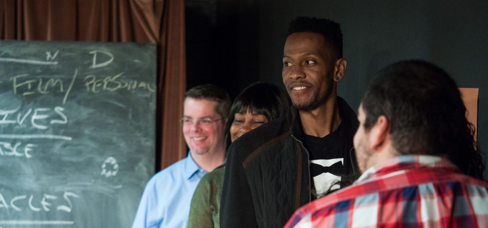A group of adults standing and smiling in a line indoors, with a chalkboard containing handwritten notes in the background.