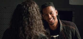 A man with short hair and earrings smiles while talking to a person with long, curly hair, seen from behind, in a dimly lit room.