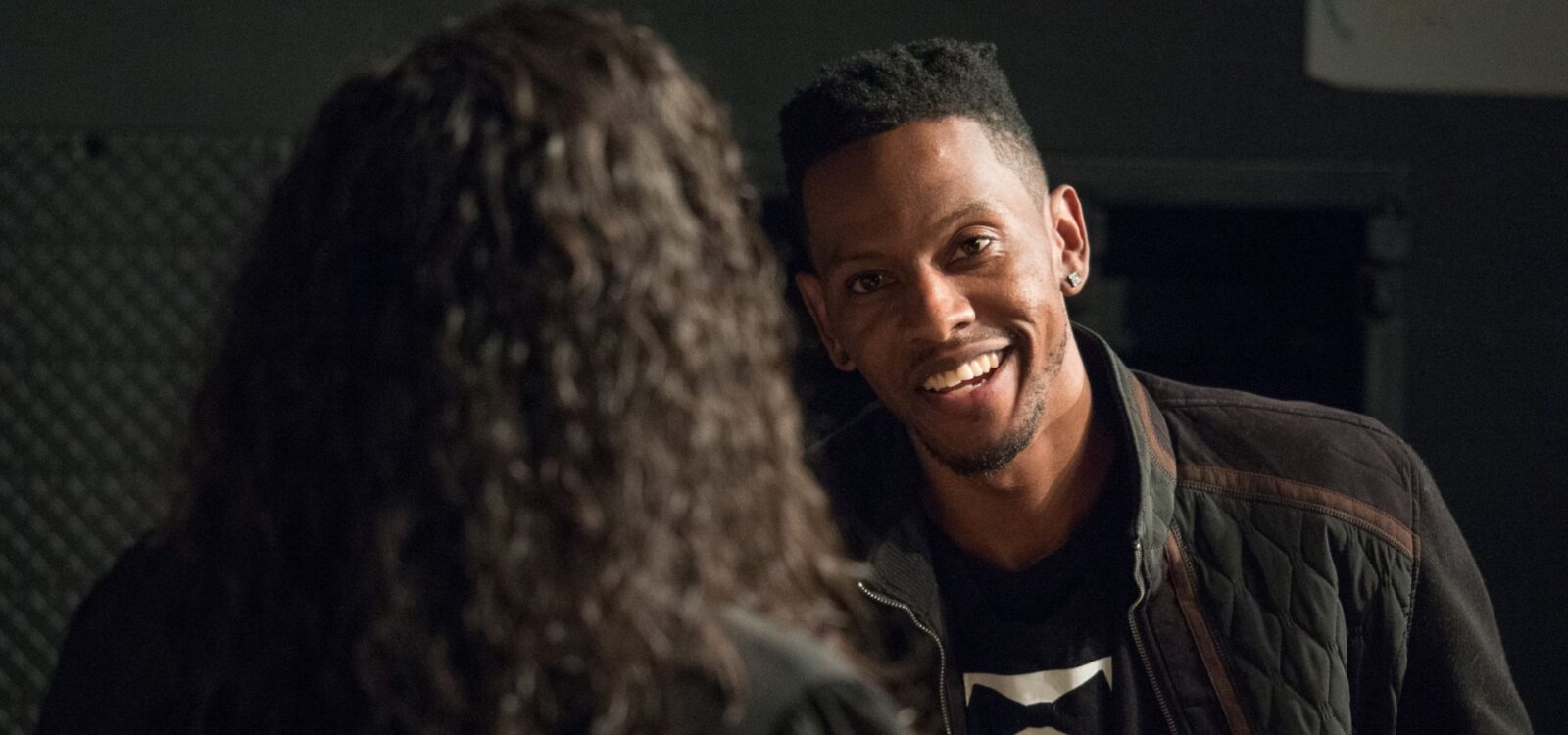 A man with short hair and earrings smiles while talking to a person with long, curly hair, seen from behind, in a dimly lit room.