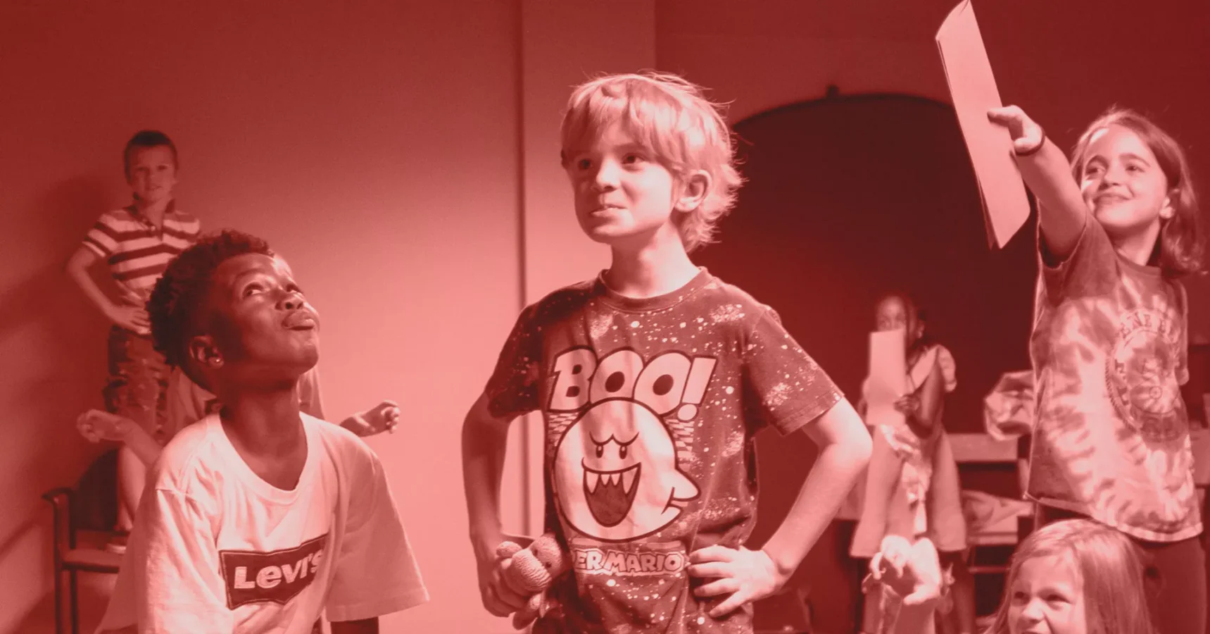 Children in a room interacting, some standing and holding papers, with a focus on two boys in the foreground, one wearing a “BOO!” shirt.