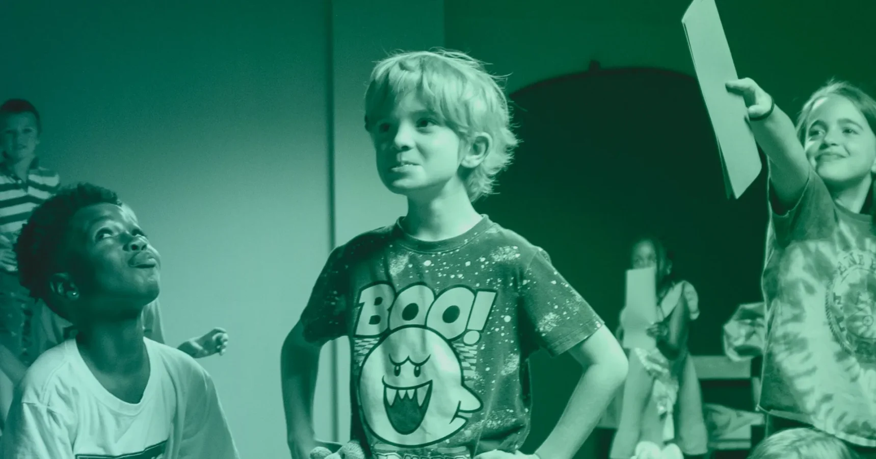 A young boy wearing a "BOO!" shirt stands with hands on hips among other children in a room, with some children smiling and holding up paper.
