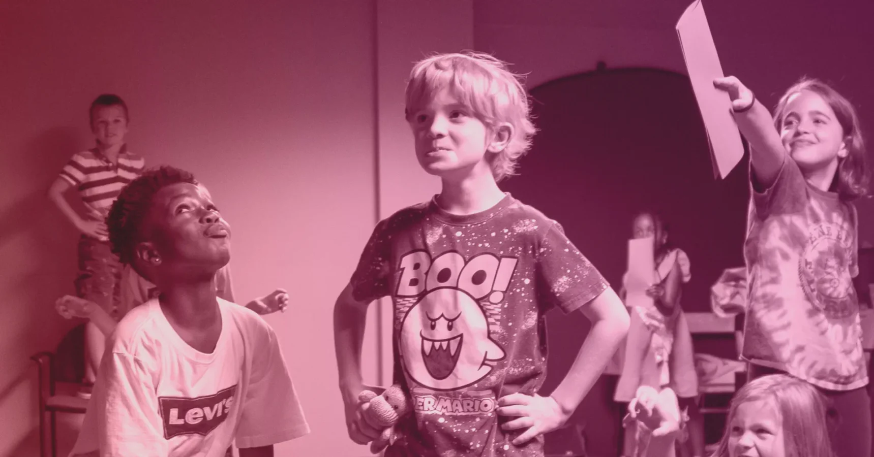 A group of children indoors, with one boy standing in the center wearing a "Boo!" shirt; others are looking at him and holding papers.