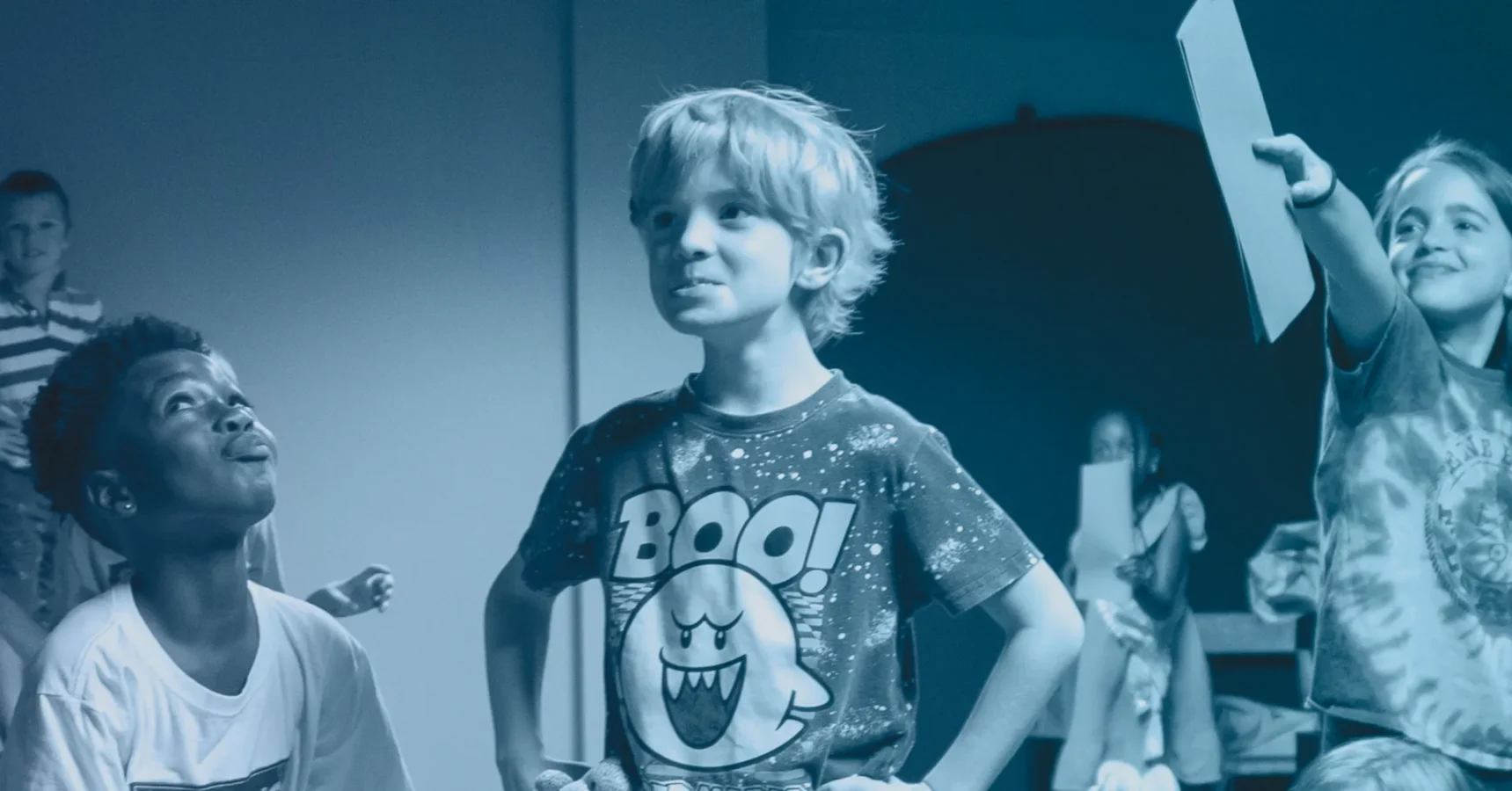 Young boy with hands on hips wearing a “Boo!” shirt stands among children indoors; other kids in the background hold up papers and smile.