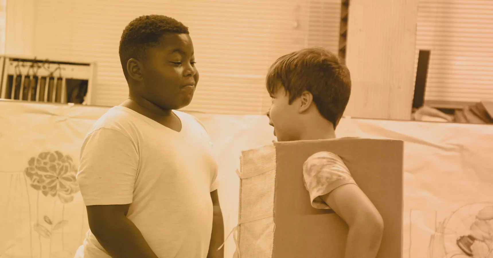 Two children interact indoors; one stands wearing a plain t-shirt, while the other faces him, partially inside a large cardboard box.