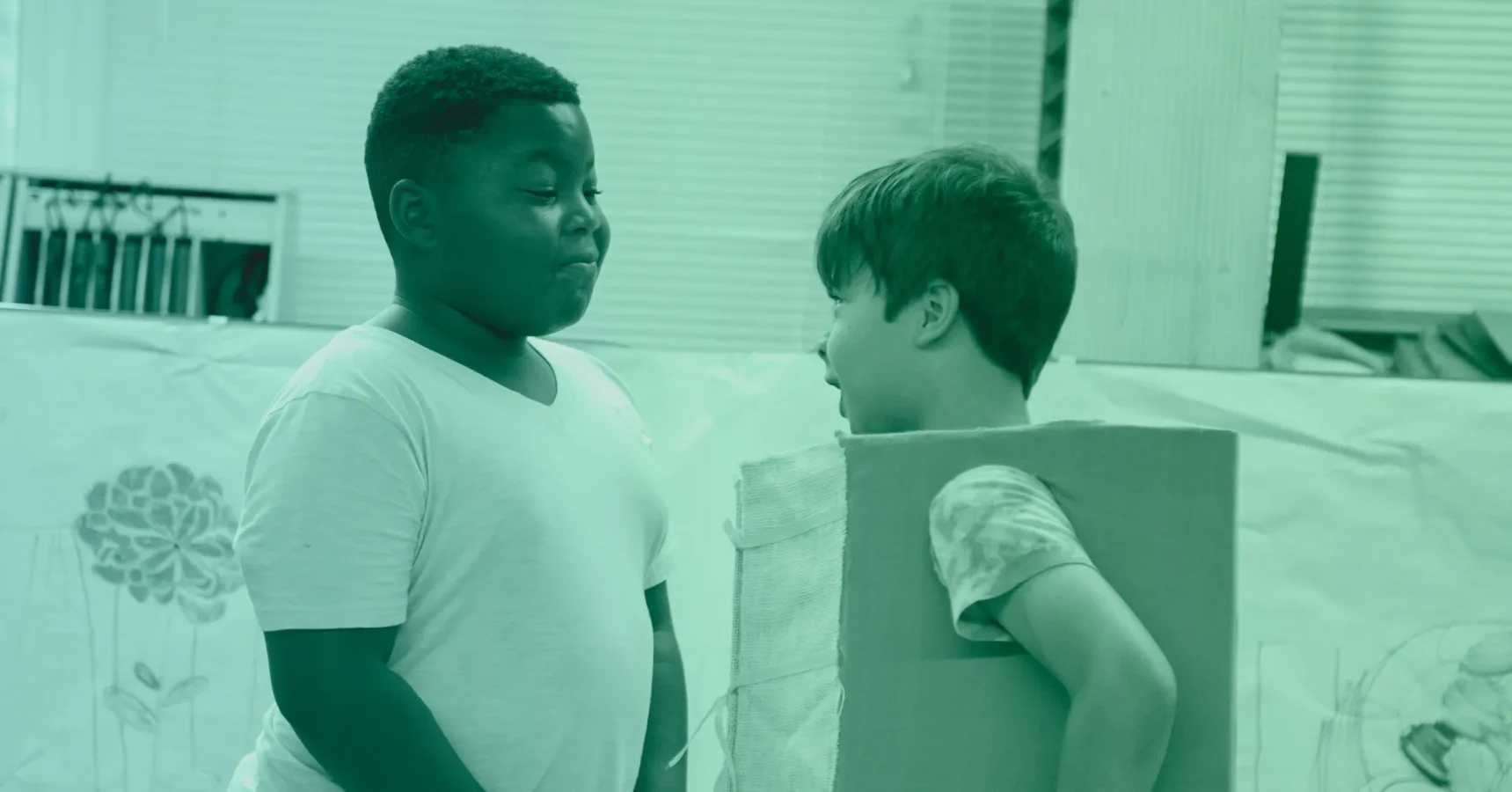 Two boys stand indoors; one faces forward in a white shirt, while the other, wearing a cardboard box, faces him and speaks.