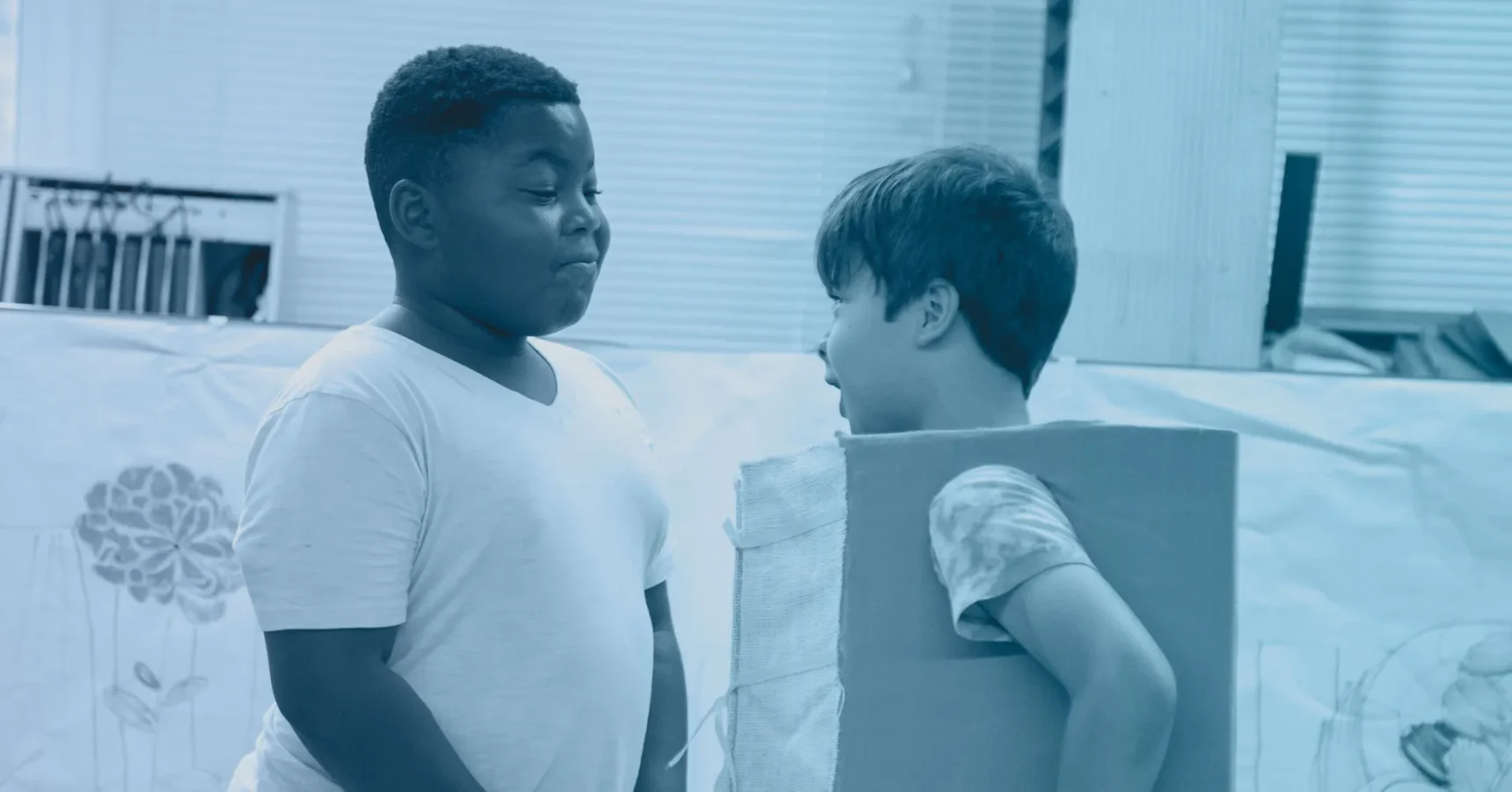 Two boys stand indoors; one wears a plain t-shirt while the other has a cardboard box around his torso. They appear to be talking or playing together.