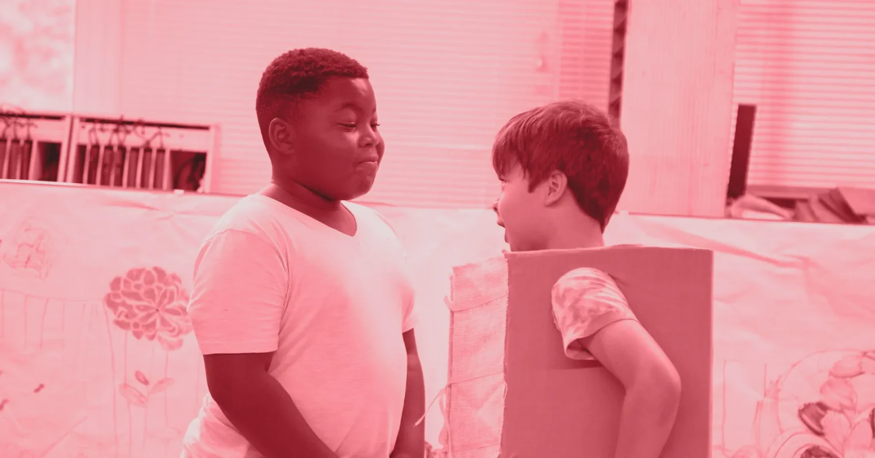 Two children stand facing each other; one wears a plain white shirt, and the other has a cardboard box around their torso. The scene appears to be indoors.