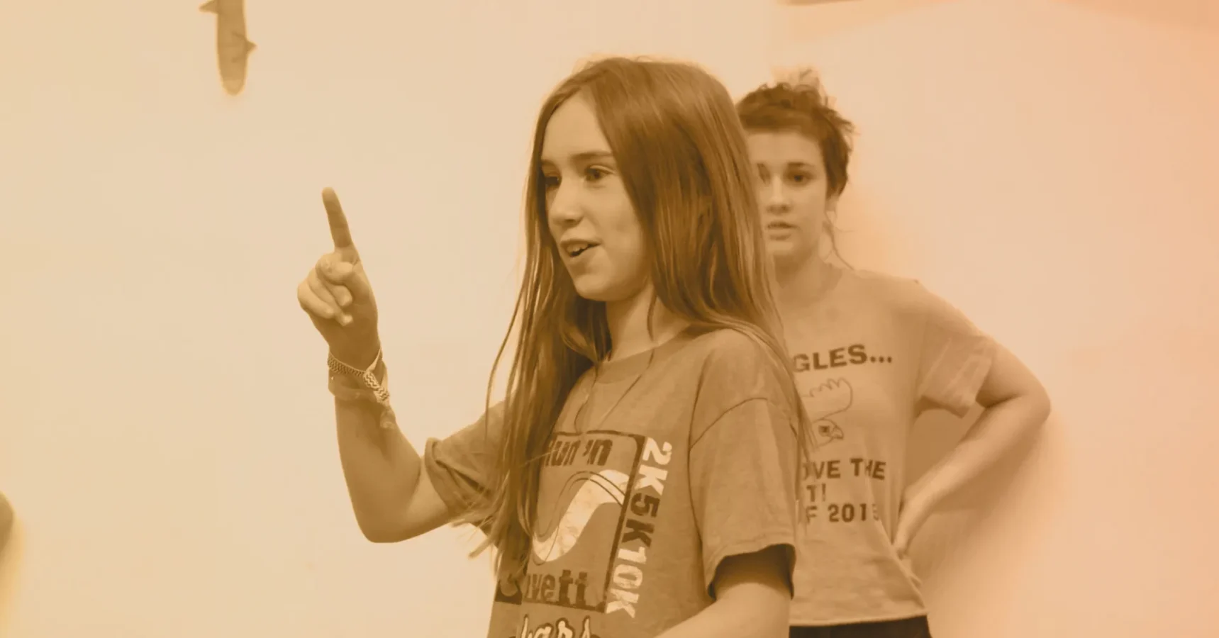 A girl with long hair gestures with her right index finger raised while another girl stands in the background, both wearing casual t-shirts.