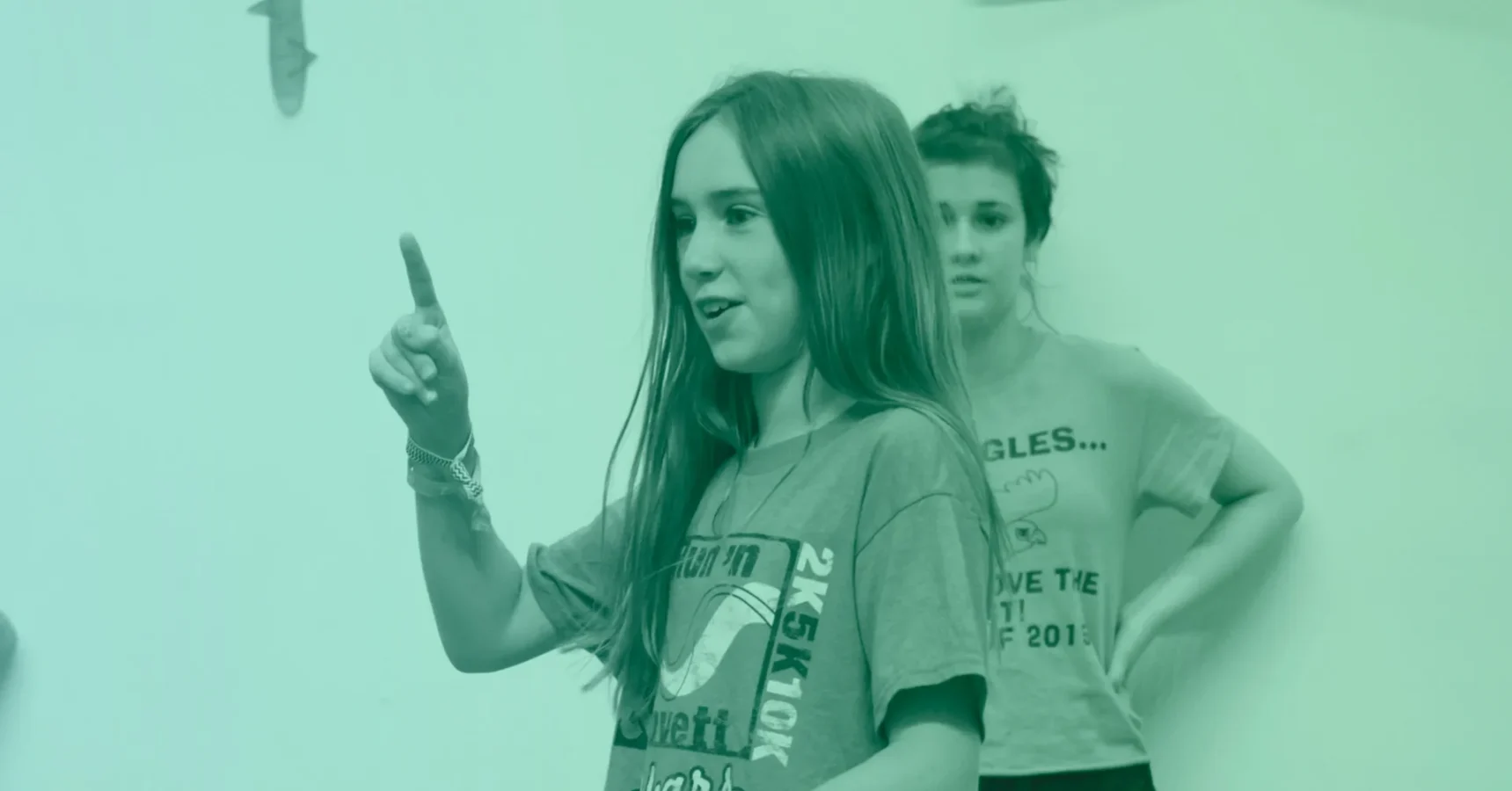 A girl stands with her right index finger raised while another girl stands behind her, both wearing casual t-shirts in a plain indoor setting.