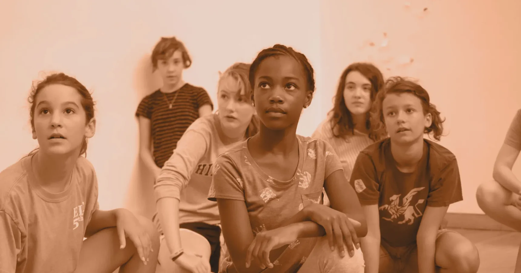 Six children sit and kneel on the floor indoors, looking attentively in various directions.