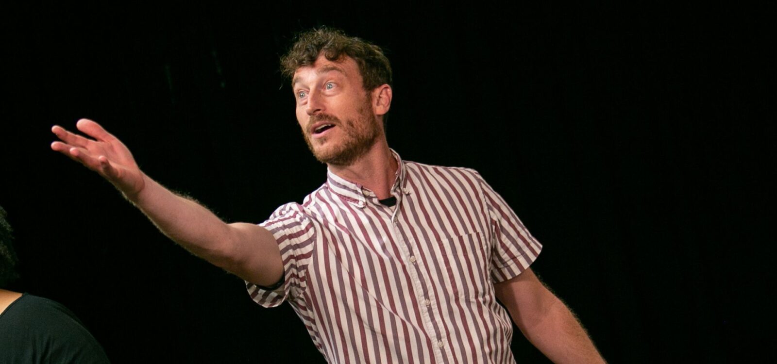 A man in a striped short-sleeve shirt stands against a black background, extending his arm and looking to the side with an expressive face.