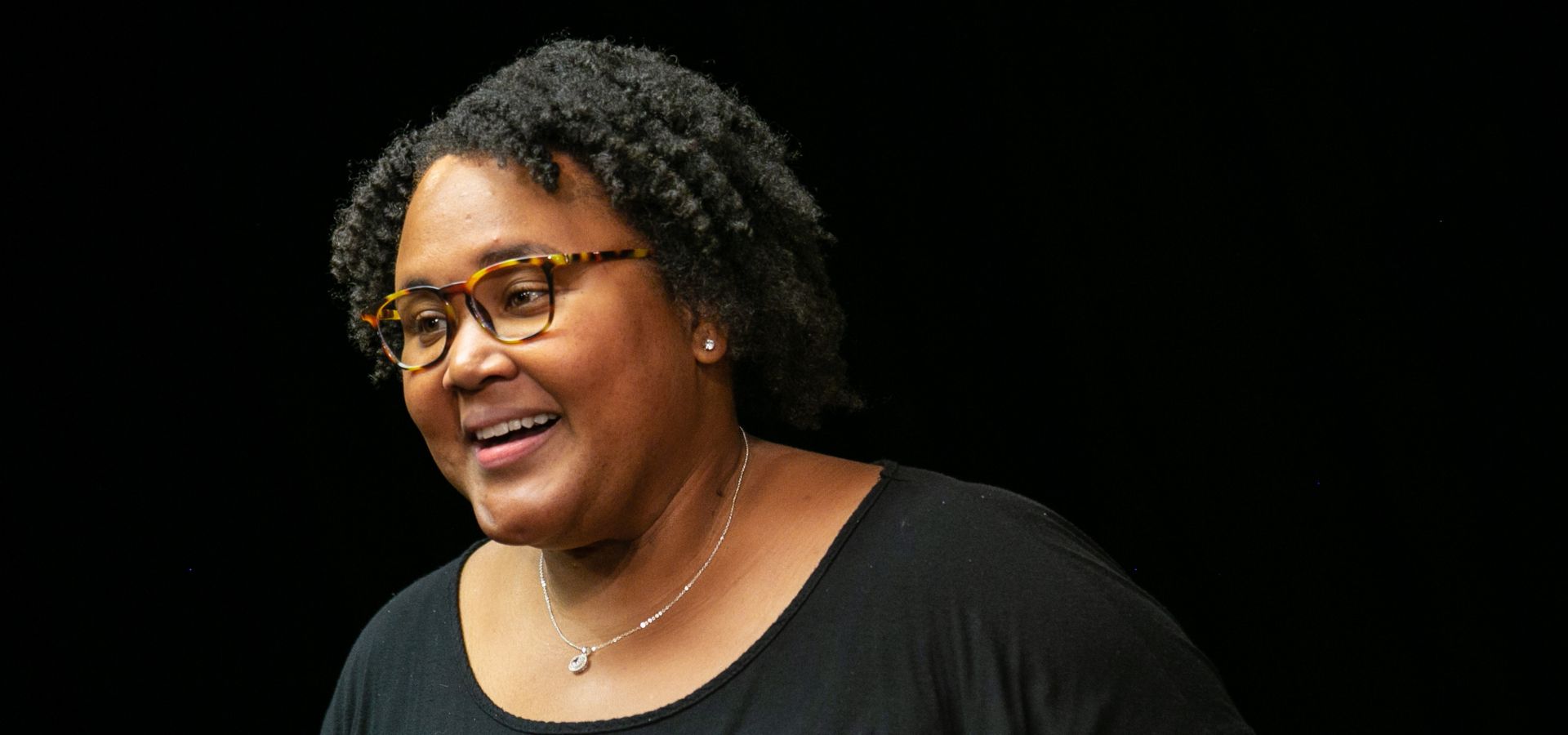 A person with short curly hair and glasses smiles, wearing a black top and a necklace, pictured against a plain black background.
