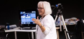 An older woman with white hair and glasses speaks in front of a camera and monitor in a room with various equipment and supplies.