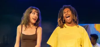 Two people stand on stage under blue lighting, both with mouths open as if singing or speaking; one wears a brown tank top, the other a yellow shirt.